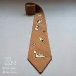 جزئیات کراوات فانتزی لینن یونیسکس به رنگ بیسکوئیتی و با طرح دایناسورهای فانتزی کوچک، چاپ شده