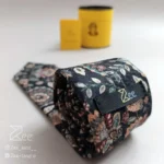 نمای پشت کراوات مخمل خیال با زمینه ی سبز تیره و طرح بوته های سنتی کرمی، با تگ برند زی همراه با جعبه استوانه فلزی نگهدارنده کراوات و دفترچه راهنمای نگهداری