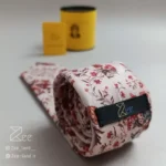 نمای پشت کراوات مخمل دیبا با زمینه ی کرمی روشن و طرح گل های ریز زرشکی و سبز، با تگ برند زی همراه با جعبه استوانه فلزی نگهدارنده کراوات و دفترچه راهنمای نگهداری